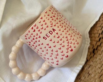 May include: A ceramic mug with a light pink exterior covered in small red hearts. The word "SEDA" is printed in red on the side. The handle is a series of connected, rounded shapes. The mug rests on a white cloth.