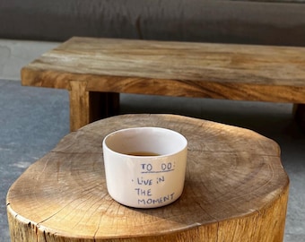 May include: A white ceramic mug with the text "TO DO: LIVE IN THE MOMENT" written on it. The mug is sitting on a wooden stump.