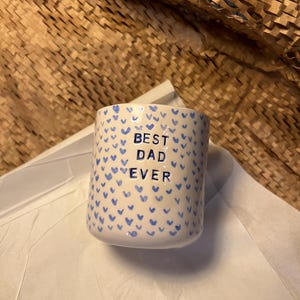 Puede incluir: Taza de cerámica blanca con un patrón de corazones azules y el texto "BEST DAD EVER". La taza es cilíndrica. El fondo incluye papel blanco y material de embalaje marrón.