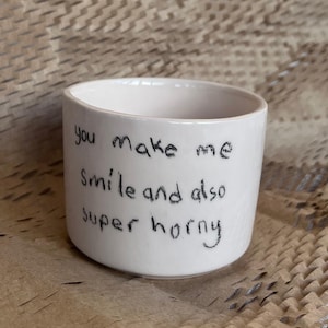 May include: A small, off-white ceramic cup with black handwritten text that reads "you make me smile and also super horny." The cup is cylindrical and sits on a brown, textured surface.