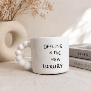 May include: A white ceramic mug with a unique bubble-textured handle. The front of the mug features the black printed text "OFFLINE IS THE NEW LUXURY" in a hand-drawn style font. The mug has a smooth, glossy finish and a cylindrical body shape.