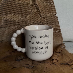 Puede incluir: Taza de cerámica blanca con asa de cuentas. La taza tiene un texto escrito a mano en negro que dice "you make me the best version of MYSELF".