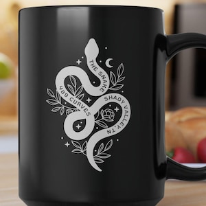 May include: Black ceramic mug featuring a white snake design with the text "The Snake 489 Curves Shady Valley TN". The mug has a large handle and is suitable for hot or cold beverages.