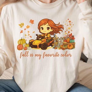 May include: Cream long-sleeved shirt with a fall-themed graphic. The graphic shows a cartoon character driving a yellow vehicle, surrounded by pumpkins and autumn leaves. The text on the shirt reads "fall is my favorite colour."