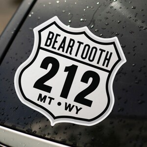 May include: A white and black sticker in the shape of a road sign, reading "BEARTOOTH 212 MT WY". The sticker is affixed to a dark, wet surface, likely a car. The design is a classic road sign style.
