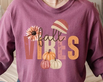 May include: A long-sleeved shirt in a maroon colour featuring a graphic design of a flower, a beanie, and pumpkins with the text "fall vibes".