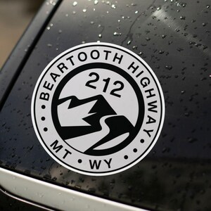 May include: A circular white sticker with a black border, featuring the text "BEARTOOTH HIGHWAY" around the edge. Inside, it has "212" above a mountain and road graphic, with "MT WY" at the bottom. The sticker is affixed to a dark, wet surface.