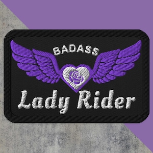 May include: Embroidered patch with the words "BADASS Lady Rider" in white thread. The design features purple wings, a heart with a rose, and a black background. The patch is rectangular with a black border.