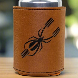 May include: A brown leather can cooler with a black spider design. The spider has eight legs and is in a web-like pattern.