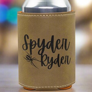 Może przedstawiać: Brązowy sk&oacute;rzany ochraniacz na puszkę z czarnym pająkiem i napisem "Spyder Ryden" w kolorze czarnym.