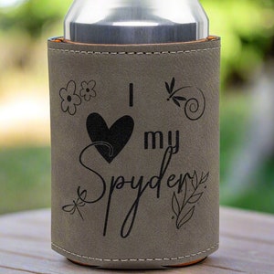 May include: Gray can cooler with a brown leatherette finish. It has a black heart with the text "I love my Spyder" and floral designs.