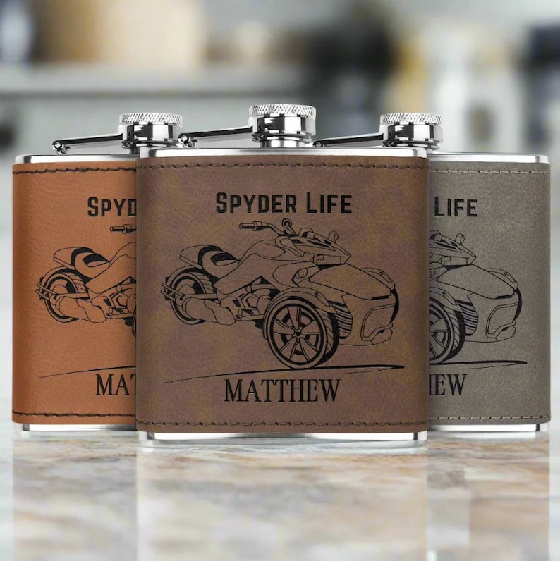 Pu&ograve; includere: Tre fiaschette in acciaio inossidabile rivestite in pelle marrone con un'illustrazione in bianco e nero di una motocicletta a tre ruote. Le fiaschette sono incise con il testo "Spyder Life", "Matthew" e "Life".