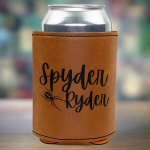 Może przedstawiać: Brązowy sk&oacute;rzany ochraniacz na puszkę z nadrukowanym czarnym pająkiem i tekstem "Spyder Ryden".