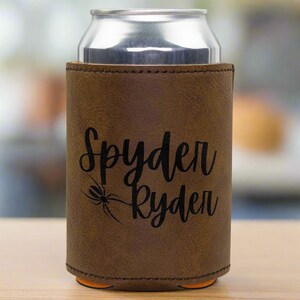 Może przedstawiać: Brązowy sk&oacute;rzany ochładzacz do puszek z czarnym pająkiem i wygrawerowanym tekstem "Spyder Ryder".
