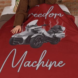 May include: A red blanket featuring a silver and black three-wheeled motorcycle design. The blanket has the words "freedom" and "Machine" in a white script font. The blanket is draped over a bed.