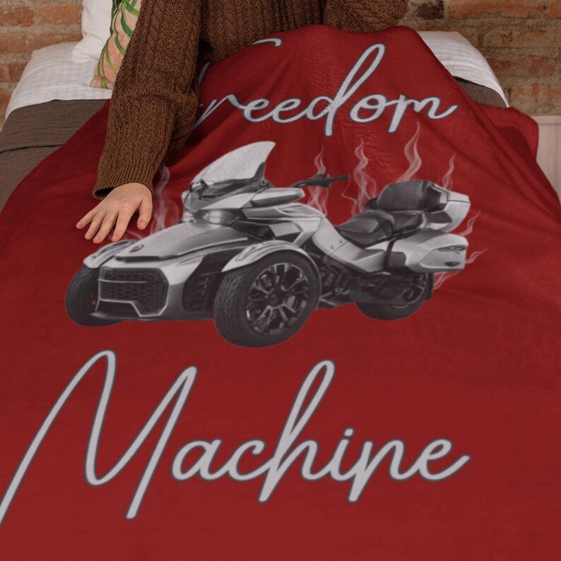 May include: A red blanket featuring a silver and black three-wheeled motorcycle design. The blanket has the words "freedom" and "Machine" in a white script font. The blanket is draped over a bed.