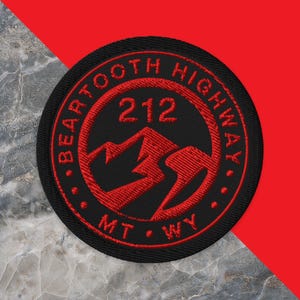 May include: A circular embroidered patch featuring a black background with red stitching. The design includes a mountain silhouette, the text "BEARTOOTH HIGHWAY", the number "212", and the letters "MT" and "WY" arranged in a circular border.