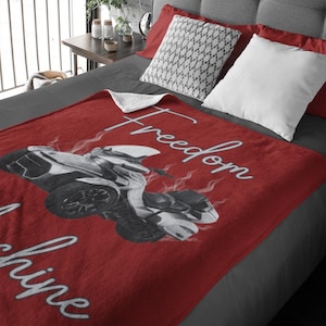 May include: A red blanket featuring a black and white motorcycle graphic with the word "Freedom" in white script. The blanket is on a bed with pillows and a side table.