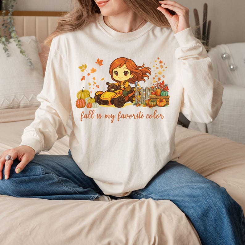 May include: Cream long-sleeved shirt with a fall-themed graphic. The graphic shows a cartoon character driving a yellow vehicle, surrounded by pumpkins and autumn leaves. The text on the shirt reads "fall is my favorite colour."