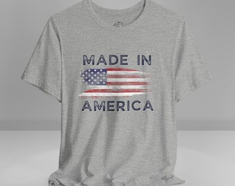 May include: A heather gray t-shirt featuring the text "MADE IN AMERICA" above and below a distressed American flag design in red, white, and blue. The shirt is made of a soft material.