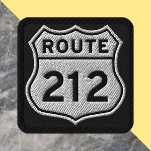 May include: Embroidered patch featuring a white and black Route 212 sign. The sign is in a shield shape with the text "ROUTE" above the number "212". The patch has a black border and a textured appearance.