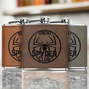 Op de afbeelding: Drie bruine, met leer beklede, roestvrijstalen heupflessen met een zwart spinnenlogo en de tekst "SPYDER CLUB". De middelste fles heeft de naam "BRENT" gegraveerd boven het logo.