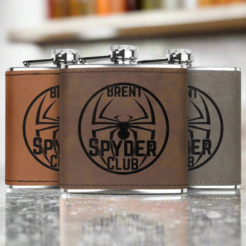 Op de afbeelding: Drie bruine, met leer beklede, roestvrijstalen heupflessen met een zwart spinnenlogo en de tekst "SPYDER CLUB". De middelste fles heeft de naam "BRENT" gegraveerd boven het logo.