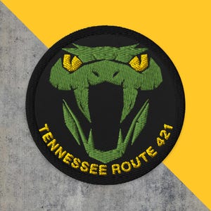 May include: Embroidered patch featuring a green snake head with yellow eyes and an open mouth on a black circular background. The text "TENNESSEE ROUTE 421" is in yellow around the bottom edge. The patch is likely for apparel or accessories.