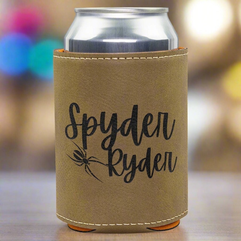 Może przedstawiać: Brązowy sk&oacute;rzany ochraniacz na puszkę z czarnym pająkiem i napisem "Spyder Ryden" w kolorze czarnym.