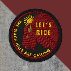 May include: Embroidered patch with a black border, featuring a red bison silhouette and a yellow sun. The text "LET'S RIDE" and "THE BLACK HILLS ARE CALLING" are in yellow. The patch is circular and has a textured appearance.