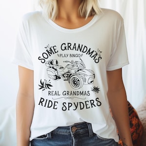 May include: White t-shirt with black text that reads "Some Grandmas Play Bingo Real Grandmas Ride Spyders". The text is surrounded by a floral design. A black and white illustration of a three-wheeled motorcycle is in the center of the shirt.