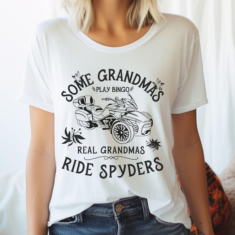 May include: White t-shirt with black text that reads "Some Grandmas Play Bingo Real Grandmas Ride Spyders". The text is surrounded by a floral design. A black and white illustration of a three-wheeled motorcycle is in the center of the shirt.
