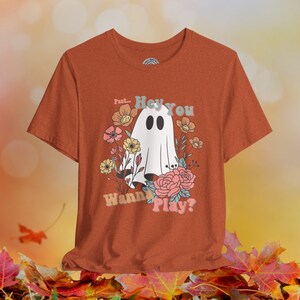 May include: Orange t-shirt with a white ghost and floral design. The text on the shirt reads "Psst... Hey You Wanna Play?"