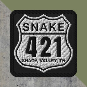 May include: Embroidered patch featuring a white and black design. The patch is in the shape of a road sign, with the text "SNAKE 421 SHADY, VALLEY, TN" in white lettering. The patch has a black border and is on a textured background.