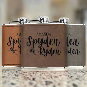 May include: Three personalized flasks in brown, dark brown, and gray leather. Each flask has a silver cap and is engraved with the name "Amanda Spyder Ryder" and a spider graphic. The flasks are displayed on a light-colored surface.