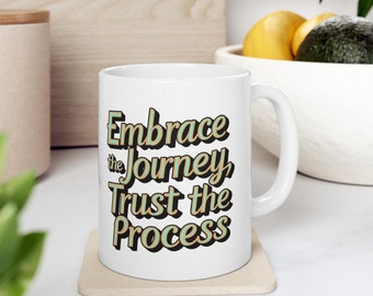 May include: A white ceramic mug with a handle, featuring the text "Embrace the Journey, Trust the Process" printed in a stylized, layered font with light blue, tan, and black colors. The mug sits on a square, light-colored coaster.