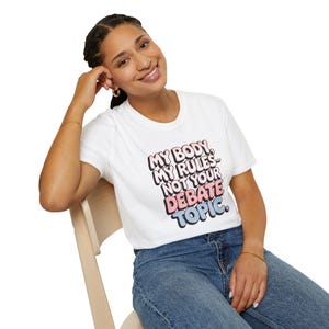 Puede incluir: Una camiseta blanca con el texto "My body, my rules, not your debate topic." en letras rosa y azul.