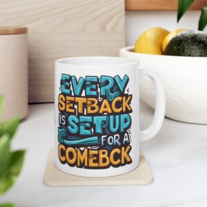 Könnte beinhalten: Weiße Keramik-Tasse mit dem Text "EVERY SETBACK IS SETUP FOR A COMEBCK" in Blau, Orange und Schwarz. Die Tasse hat einen gebogenen Henkel und steht auf einem hellen Untersetzer. Die Tasse steht auf einer weißen Oberfläche.