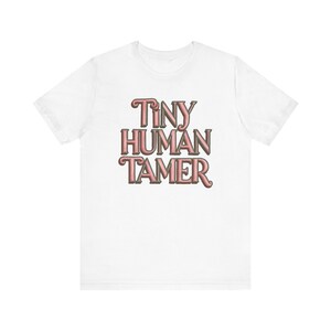 Può includere: Una t-shirt bianca a maniche corte con la scritta "TINY HUMAN TAMER" stampata in un carattere rosa stilizzato e stratificato con un contorno scuro centrato sul petto. Il capo presenta uno scollo rotondo e una vestibilità standard.