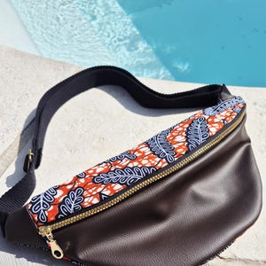 May include: A brown fanny pack with a patterned top section featuring orange, white, and navy blue leaf designs. The bag has a gold zipper and a black adjustable strap. The fanny pack is made of leather-like material.
