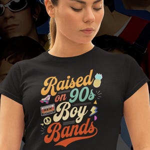 Pode incluir: Camiseta preta com o texto "Raised on 90s Boy Bands" em amarelo, laranja e azul. O design inclui um patins, um abacaxi, uma fita cassete, um símbolo da paz e um raio. A camiseta é usada por uma pessoa.