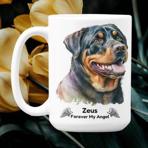 Puede incluir: Taza de cerámica blanca con un retrato de perro Rottweiler en negro, marrón y canela. La taza tiene el texto "Zeus Forever My Angel" con alas de ángel. El fondo incluye flores amarillas y follaje verde oscuro.