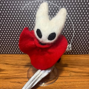 May include: A handmade felt figure of a character with a white head, black eyes, and a red cape. The figure is holding a silver-colored sword and is mounted on a clear stand. The background is a black and white polka dot pattern.