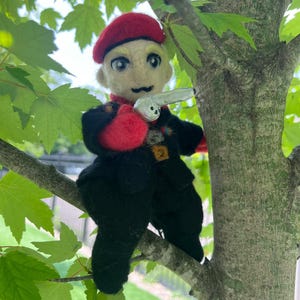Może przedstawiać: Ręcznie wykonana filcowa lalka przedstawiająca osobę w czerwonym berecie i czarnym mundurze, trzymającą srebrny pistolet-zabawkę. Lalka siedzi na gałęzi drzewa z zielonymi liśćmi w tle. Twarz lalki ma neutralny wyraz.