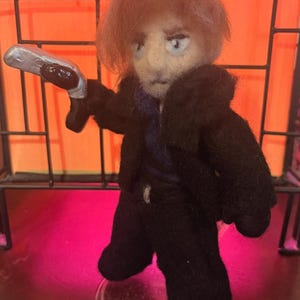 May include: A needle-felted doll with light brown hair, wearing a black coat over a dark purple shirt and black trousers. The doll has a serious facial expression and is holding a silver-colored curved blade in its right hand, standing on a clear base.