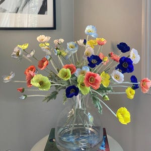 Op de afbeelding: Een boeket kleurrijke zijden papaverbloemen in een heldere glazen vaas. De bloemen zijn in tinten geel, oranje, roze, blauw en groen.