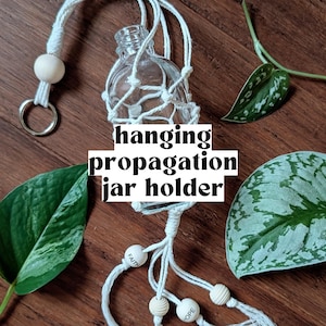 Op de afbeelding: Een heldere glazen propagatiepot die is opgehangen in een witte macraméhouder. De houder heeft houten kralen en de woorden "Faith" en "Hope". De tekst "hanging propagation jar holder" is over de afbeelding geplaatst. Groene bladeren zijn zichtbaar.