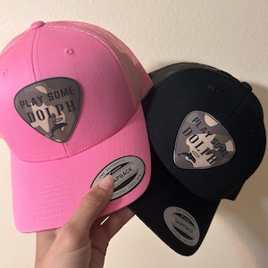 Puede incluir: Se muestran dos gorras de camionero, una rosa y otra negra. Cada gorra tiene una visera curva, una parte trasera de malla y un parche con el texto "PLAY SOME DOLPH" en blanco. La gorra rosa tiene una visera rosa a juego.