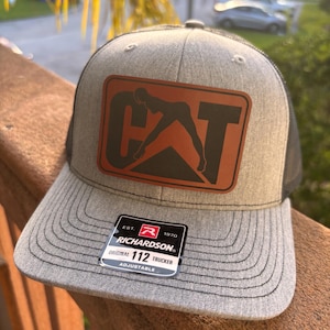 Op de afbeelding: Een grijze truckerpet met een zwarte mesh achterkant en een gebogen klep. De pet heeft een bruine leren patch met het woord "CAT" en een silhouet van een persoon. Een zwart-wit label is aan de klep bevestigd.