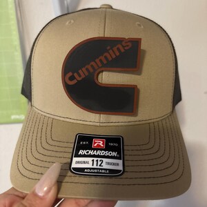 May include: Tan and black trucker hat with the Cummins logo in black and brown. The hat has a curved brim and a mesh back. The Richardson tag is visible on the front. The hat is adjustable.